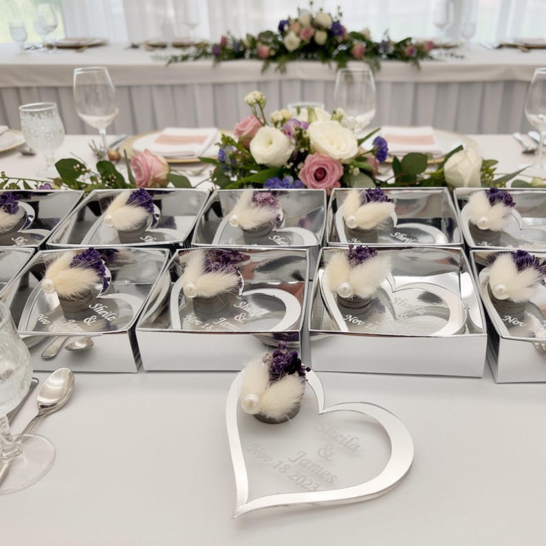 May include: A collection of silver heart-shaped acrylic frames, each featuring a small cluster of dried purple flowers and white pampas grass with a pearl accent. The frames are engraved with the text Sheila & James Nov 18 2023.