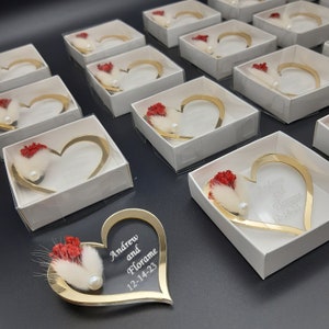 May include: Gold heart-shaped boxes with a red flower and white feather inside. The boxes are personalized with the names "Andrew and Florame" and the date "12-14-23".