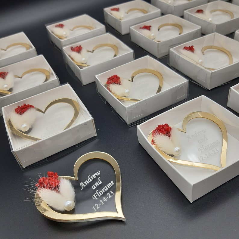 May include: Gold heart-shaped boxes with a red flower and white feather inside. The boxes are personalized with the names "Andrew and Florame" and the date "12-14-23".