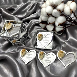 May include: Heart-shaped silver mirror favors with the text Thank You for Celebrating with Us, each featuring a small beige dried flower and a white pearl accent. Some mirrors are inside square silver boxes, accompanied by a cotton stem.