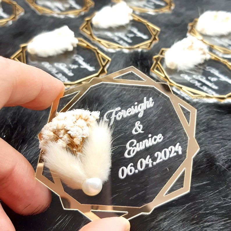 May include: A clear acrylic octagon-shaped wedding favor with a gold border. The favor features a dried flower arrangement and a pearl. The text "Foresight & Eunice 06.04.2024" is engraved on the acrylic.