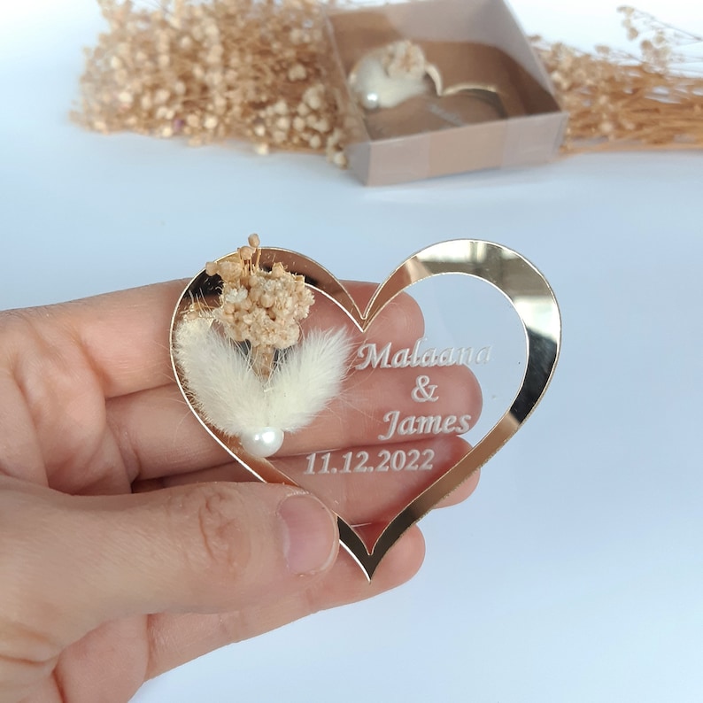 May include: A gold heart-shaped mirror with a white feather and dried flowers inside. The mirror is engraved with the names "Malaana & James" and the date "11.12.2022".