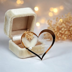 May include: A cream-colored velvet ring box paired with a heart-shaped gold-toned acrylic ornament. The ornament features dried beige flowers, a small white pearl, and white engraved text reading Malaona & James and 11.12.2022.