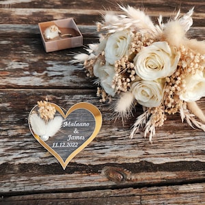 May include: A heart-shaped plaque with a gold border and dark center, featuring the white text "Malaano & James 11.12.2022" and a small white feather accent, displayed next to a bouquet of cream roses and dried grasses and a small gift box.