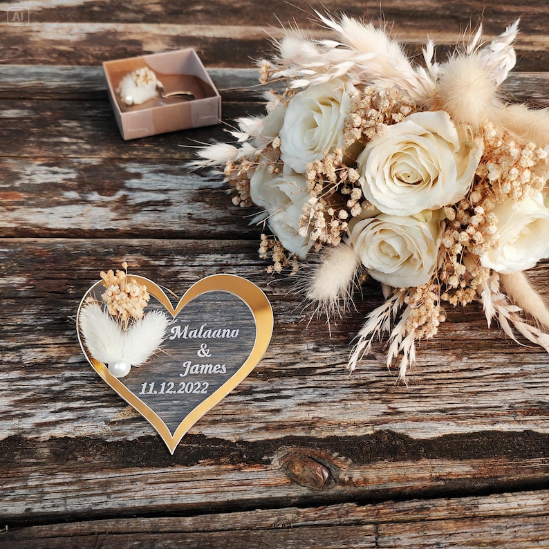 May include: A heart-shaped plaque with a gold border and dark center, featuring the white text "Malaano & James 11.12.2022" and a small white feather accent, displayed next to a bouquet of cream roses and dried grasses and a small gift box.