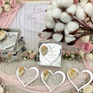 May include: A collection of heart-shaped silver mirror acrylic favors featuring the engraved text "Thank you for celebrating with us." Each piece is adorned with a small dried floral accent. The items are displayed alongside white cotton bolls.