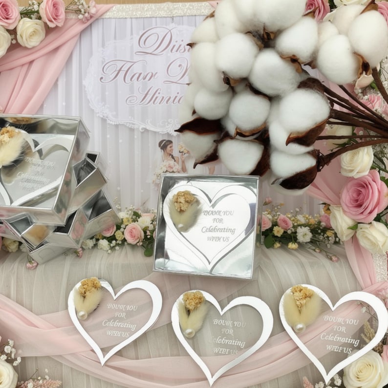 May include: A collection of heart-shaped silver mirror acrylic favors featuring the engraved text "Thank you for celebrating with us." Each piece is adorned with a small dried floral accent. The items are displayed alongside white cotton bolls.