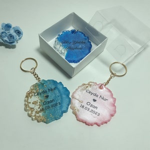 May include: Two round resin keychains with gold flakes. One is blue and white with the text "Ceyda Nur Ozan 18.03.2023". The other is pink and white with the text "Ceyda Nur Ozan 18.03.2023".