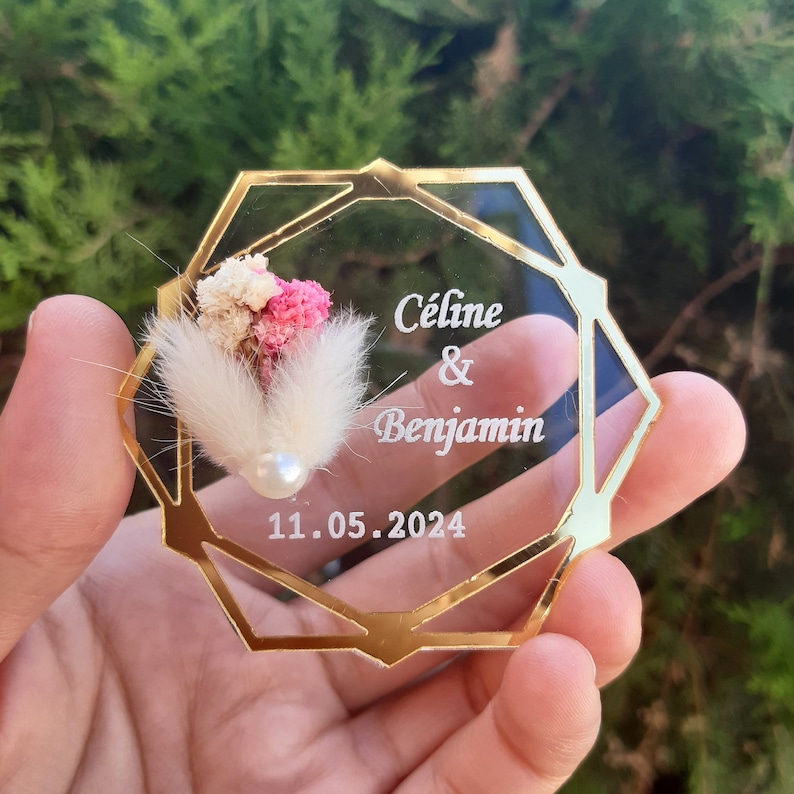 May include: A clear, octagonal, gold-rimmed acrylic favor box with a small bouquet of dried flowers and a pearl. The box is engraved with the text "C&eacute;line & Benjamin" and the date "11.05.2024".