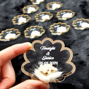 May include: A clear acrylic wedding favor with a gold border in the shape of a seashell. The favor has the names "Foresight & Eunice" and the date "06.06.2024" engraved on it. A small white flower with a pearl is attached to the favor.