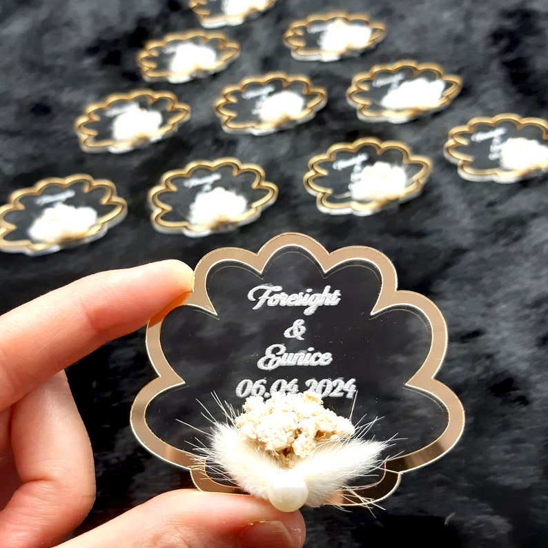 May include: A clear acrylic wedding favor with a gold border in the shape of a seashell. The favor has the names "Foresight & Eunice" and the date "06.06.2024" engraved on it. A small white flower with a pearl is attached to the favor.