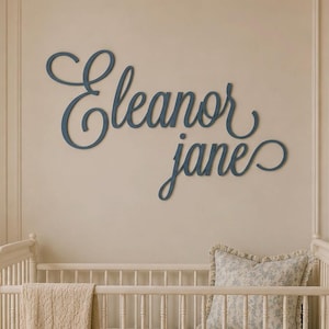 May include: A dark blue wall-mounted sign featuring the name Eleanor Jane in a cursive script font. The letters are connected and mounted directly onto the wall, displaying a smooth finish and a flowing, decorative style for interior wall decor.