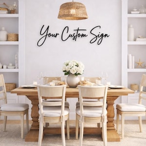 May include: A black script wall sign with the text Your Custom Sign mounted on a white wall above a wooden dining table with four chairs, a white floral centerpiece, and a woven pendant light fixture hanging from the ceiling above the table.
