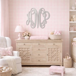 May include: A nursery with a light pink and white color scheme. A large, gray monogram hangs on the wall above a light wood dresser. A white armchair with a pink bow pillow sits to the left. A crib and shelves are on the right.