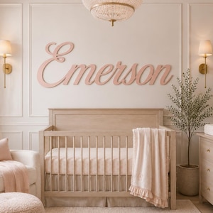 May include: A nursery room featuring a light wood crib with a soft pink blanket, a cream-colored armchair with a matching ottoman, and a wooden dresser. The wall displays the name Emerson in large, cursive, light pink wooden lettering.
