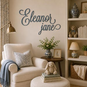 May include: A dark blue wall-mounted sign featuring the name Eleanor Jane in a cursive script font. The letters are connected in a flowing, decorative style, mounted directly onto a light-colored wall above a cream-colored armchair.