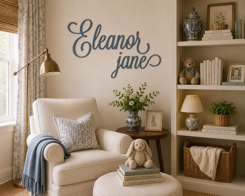 May include: A dark blue wall-mounted sign featuring the name Eleanor Jane in a cursive script font. The letters are connected in a flowing, decorative style, mounted directly onto a light-colored wall above a cream-colored armchair.