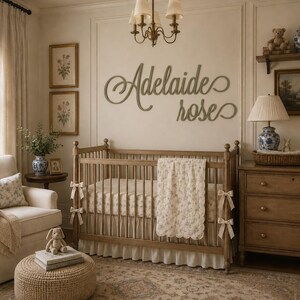 May include: A wooden wall sign featuring the name Adelaide rose in a cursive script font, mounted above a wooden crib with a white floral quilt, a beige armchair, a wooden dresser, and a lamp in a room with framed botanical wall art.