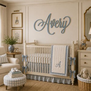 May include: A nursery room featuring a white crib with a blue and white quilt monogrammed with the letter A, a blue wooden name sign reading Avery on the wall, a cream armchair, a blue gingham ottoman, and a wicker basket with a plush rabbit.
