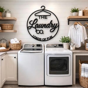 May include: A circular black wall sign featuring a hanger icon and the text "THE Laundry CO. LOADS OF FUN" mounted above two white washing machines. The laundry room includes wooden shelves, woven baskets, a white shirt, and green plants.