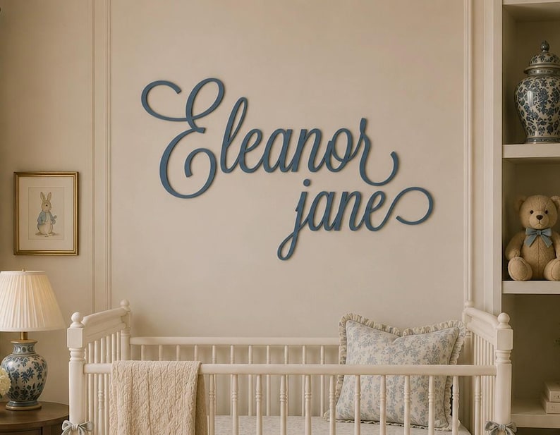 May include: A dark blue wall-mounted sign featuring the name Eleanor Jane in a cursive script font. The letters are connected and mounted directly onto the wall, displaying a smooth finish and a flowing, decorative style for interior wall decor.