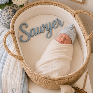 May include: A newborn baby wrapped in a white swaddle blanket and wearing a blue and white striped hat, resting in a woven wicker bassinet. A blue wooden cutout with the name Sawyer is placed inside next to the baby on the white liner.