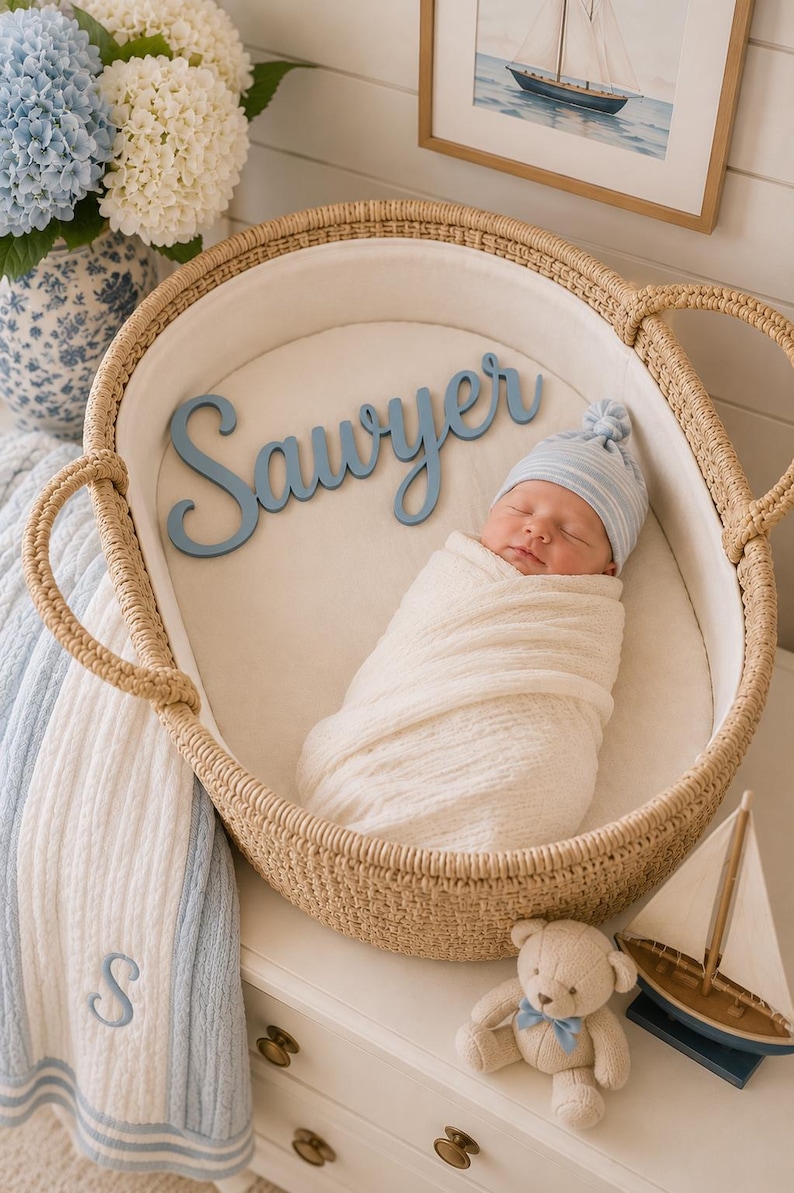 May include: A newborn baby wrapped in a white swaddle blanket and wearing a blue and white striped hat, resting in a woven wicker bassinet. A blue wooden cutout with the name Sawyer is placed inside next to the baby on the white liner.