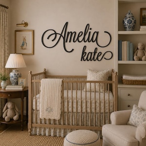 May include: A black script wall sign featuring the name Amelia Kate mounted above a wooden crib. The crib is dressed with a patterned sheet and a cream-colored quilted blanket. The room includes a side table, a lamp, and a bookshelf.