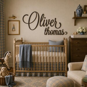 May include: A dark brown wooden wall sign featuring the name Oliver Thomas in a cursive script font, mounted above a light wood crib with a blue plaid skirt, a blue throw blanket, and a gray pillow in a nursery setting.