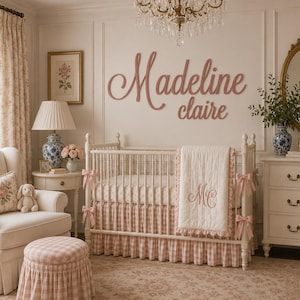 May include: A nursery room featuring a white crib with a pink and white gingham skirt and a quilt embroidered with the initials MC. Above the crib, the name Madeline claire is displayed in large, pink, cursive wooden wall lettering.
