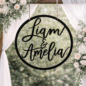 May include: A black circular sign with the names "Liam & Amelia" in elegant cursive script, suspended by twine. The sign is set against a backdrop of white flowers and greenery, suggesting a wedding or event setting.