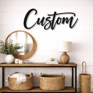 May include: A black wall-mounted sign featuring the word Custom in a cursive script font. The sign is displayed on a white shiplap wall above a wooden console table with a black metal frame, two woven baskets, a round mirror, and a table lamp.