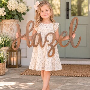 May include: A large, dark brown wooden cutout sign featuring the name Hazel in a cursive script font. The sign is crafted from smooth wood with a natural grain finish and is held horizontally by a child wearing a cream floral print dress.