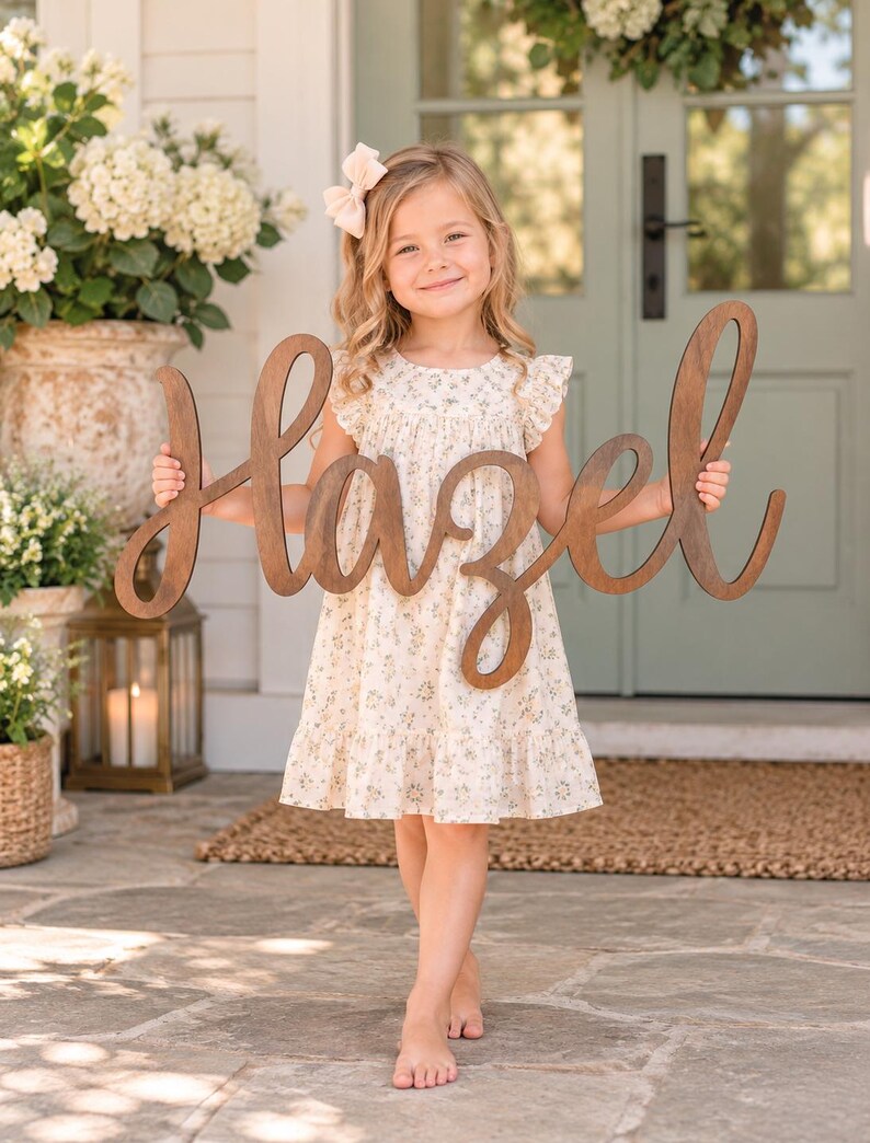 May include: A large, dark brown wooden cutout sign featuring the name Hazel in a cursive script font. The sign is crafted from smooth wood with a natural grain finish and is held horizontally by a child wearing a cream floral print dress.