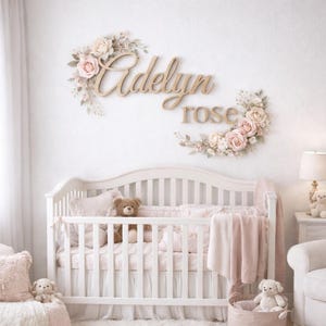 May include: A white wooden crib with a light pink blanket and teddy bears. Above the crib, a wall sign displays the name Adelyn rose in cursive wooden lettering, accented by two floral arrangements of pink and white roses and leaves.