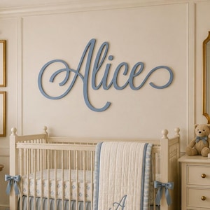 May include: A large, blue, cursive wall cutout of the name Alice, featuring elegant, flowing script lettering. The letters are connected in a single, continuous piece, mounted horizontally on a light-colored wall above a wooden crib.