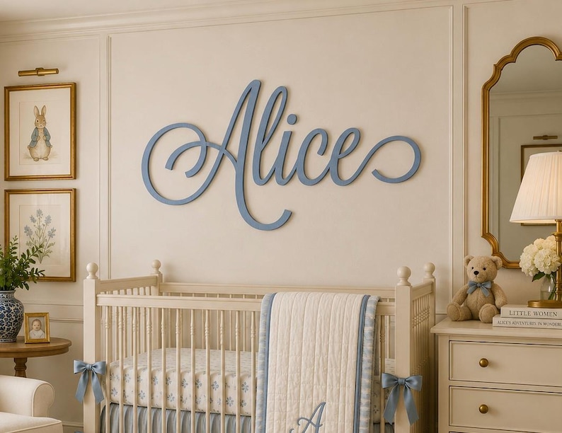 May include: A large, blue, cursive wall cutout of the name Alice, featuring elegant, flowing script lettering. The letters are connected in a single, continuous piece, mounted horizontally on a light-colored wall above a wooden crib.