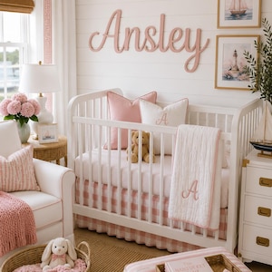 May include: A white wooden crib with a pink and white gingham bed skirt, a white quilt with a pink embroidered letter A, a pink pillow, a white pillow with a pink letter A, and a teddy bear. The wall features the name Ansley in pink.