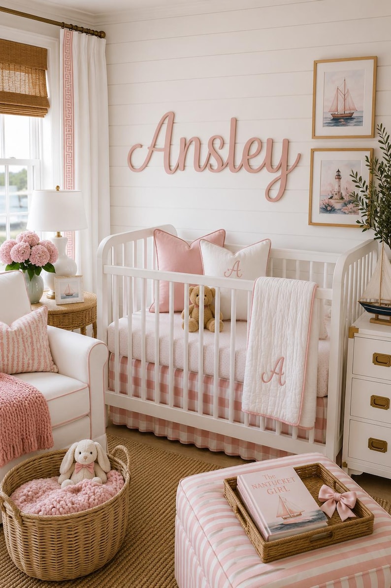 May include: A white wooden crib with a pink and white gingham bed skirt, a white quilt with a pink embroidered letter A, a pink pillow, a white pillow with a pink letter A, and a teddy bear. The wall features the name Ansley in pink.