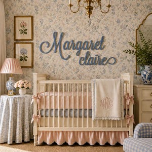 May include: A white wooden crib with a pink ruffled skirt, a pink mattress, and a cream-colored blanket featuring a monogram. Above the crib, blue cursive wooden lettering displays the name Margaret Claire on a floral wallpaper background.