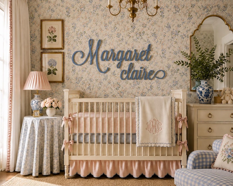 May include: A white wooden crib with a pink ruffled skirt, a pink mattress, and a cream-colored blanket featuring a monogram. Above the crib, blue cursive wooden lettering displays the name Margaret Claire on a floral wallpaper background.