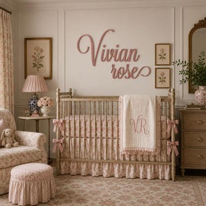 May include: A wooden crib with a light wood finish, featuring a ruffled floral skirt, a cream-colored blanket with the embroidered initials VR, and pink ribbon accents. Above the crib, the name Vivian rose is displayed in pink script lettering.