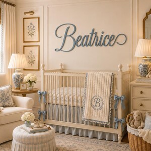 May include: A wall-mounted wooden name sign in a cursive script reading Beatrice in a muted blue finish, positioned above a cream-colored wooden crib with a cable-knit blanket featuring a circular monogram with the letter B.