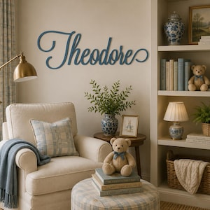 May include: A dark blue wooden wall sign featuring the name Theodore in a cursive script font. The letters are connected in a flowing style and mounted on a plain wall above a cream-colored armchair, a small wooden table, and a bookshelf.