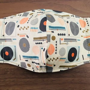 May include: A white face mask with a black elastic band. The mask has a repeating pattern of black and white vinyl records, orange and white circles, and gray and blue geometric shapes.