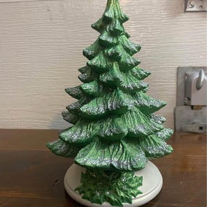 May include: A green ceramic Christmas tree with a white base and a glittery finish. The tree is decorated with holly leaves.