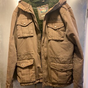 Op de afbeelding: Een bruine jas met capuchon met meerdere voorzakken en het woord "KAPORA" op de voorkant gedrukt. De jas heeft een groene, fuzzy binnenvoering en een trekkoord in de taille. De jas hangt aan een metalen hanger.