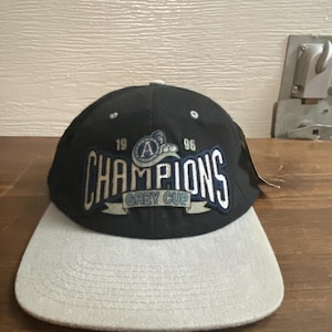 May include: A black and grey baseball cap with the word "CHAMPIONS" in white and blue lettering. The cap has a grey brim and features the text "GREY CUP" and the numbers "19" and "96".