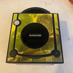 Könnte beinhalten: Eine schwarze Nintendo GameCube Spielkonsole mit einer individuellen grünen Folierung auf der Oberseite. Die Folie zeigt die Grafik einer Figur mit blondem Haar und enthält das Nintendo GameCube Logo in der Mitte der runden Disc-Abdeckung.