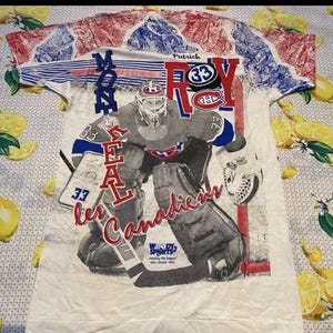 May include: White t-shirt with a hockey goalie in a Montreal Canadiens uniform. The shirt has red, white, and blue accents, with the words "Montreal" and "Canadiens" displayed. The goalie's number 33 is visible.